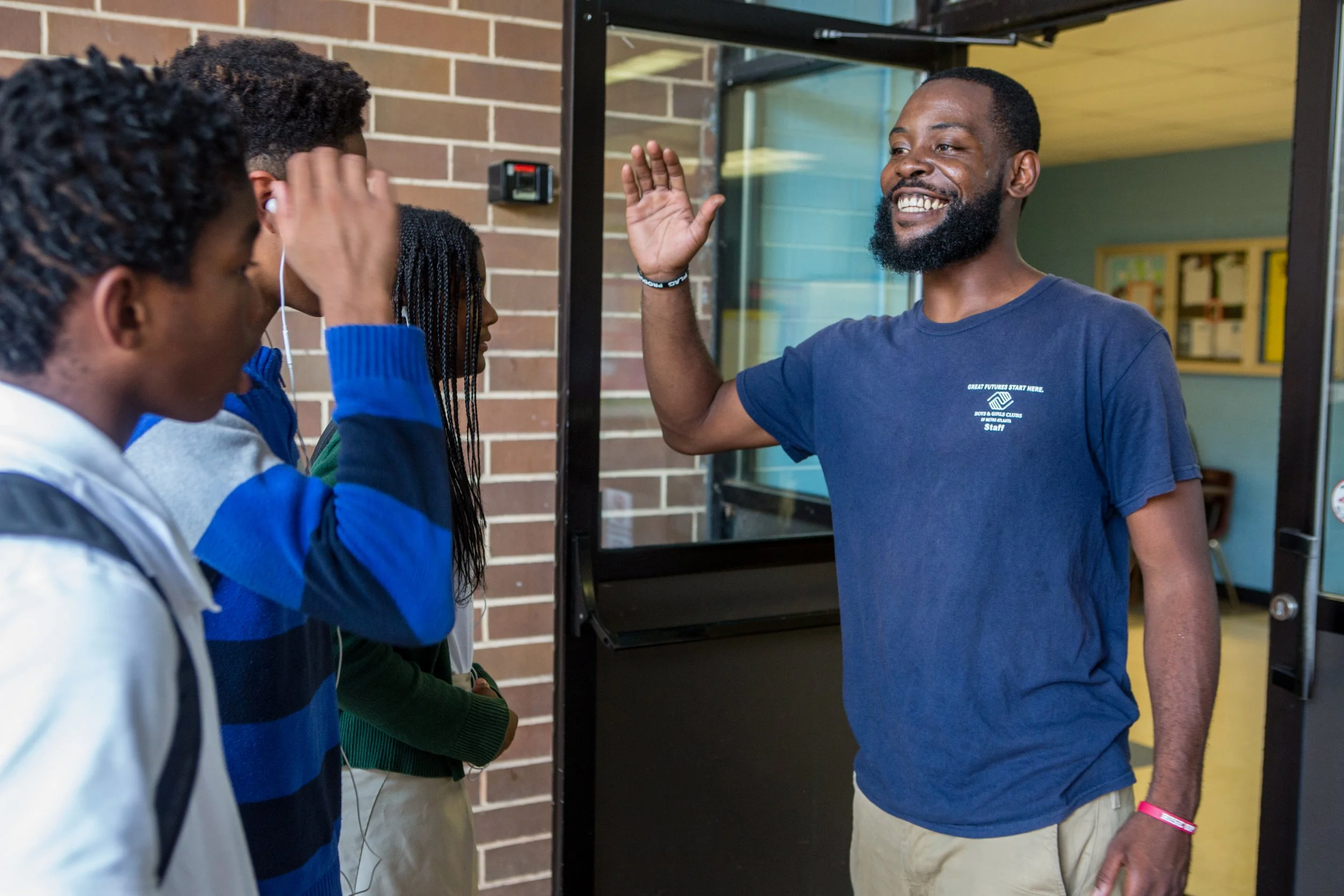 A smiling man in a blue t-shirt high-fives a group of students as they walk into a school building.