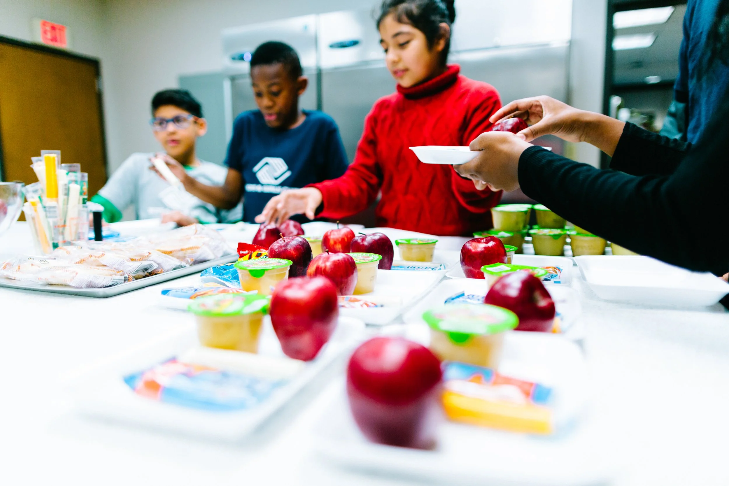 Children at a table with apples, yogurt, and snacks during a food distribution or cultural event.