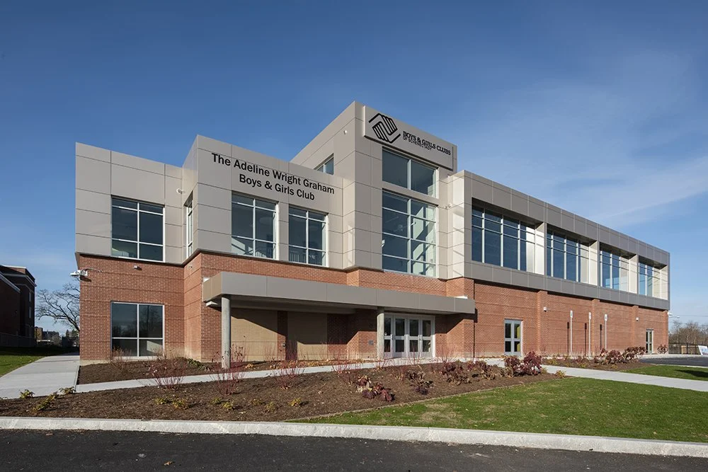Modern two-story building with brick and glass exterior, labeled as The Adeline Wright Graham Boys & Girls Club, with a blue sky background and landscaped front yard.