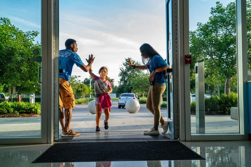 Young girl running outside as two staff members high-five her at the entrance of a building on a sunny day.