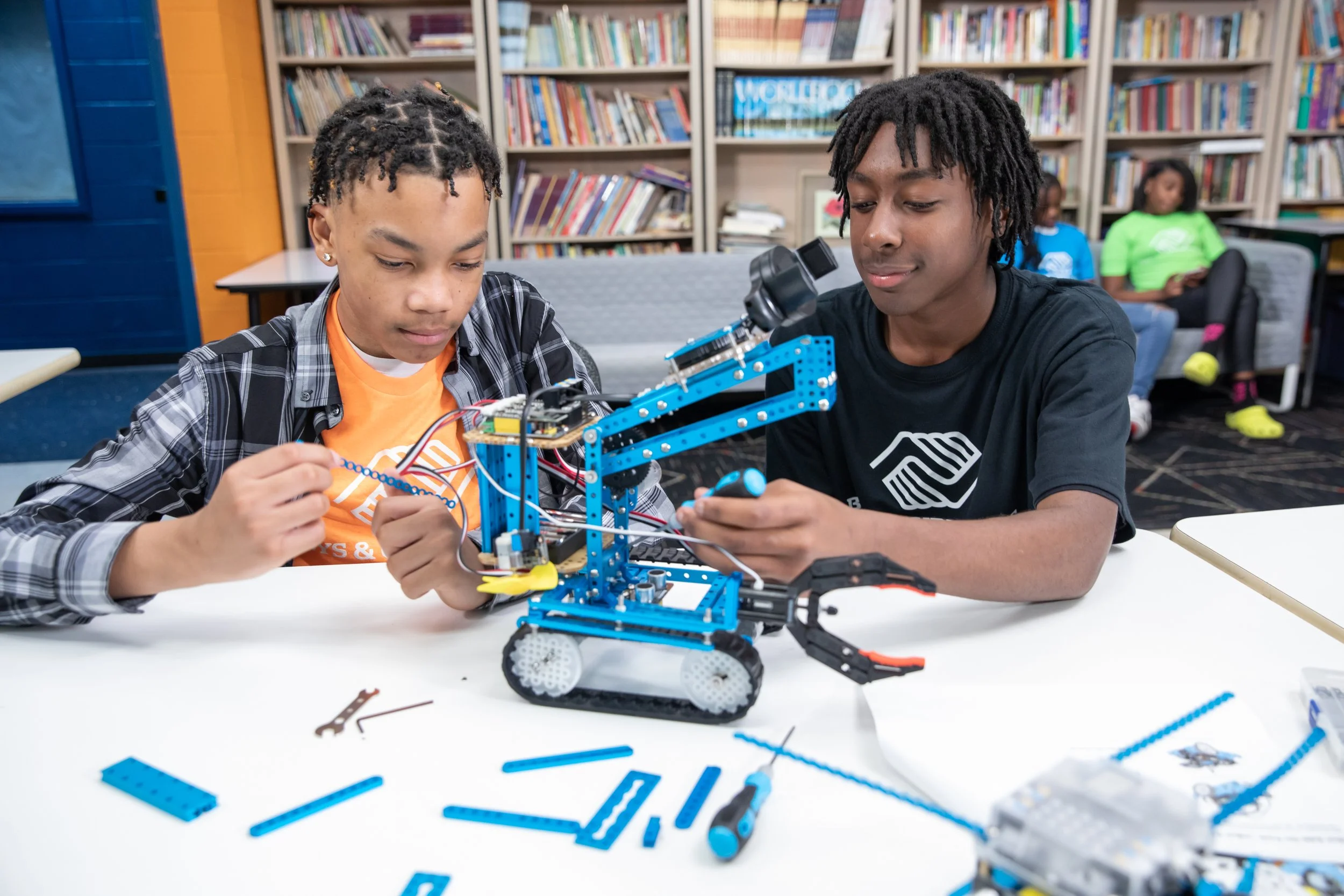 Two boys working on assembling a robot with blue and black parts at a table in a classroom or library, with a bookshelf in the background.