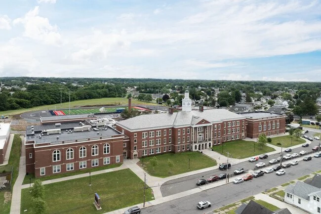 Aerial view of a brick school building with a clock tower, sports field, and parking lot.