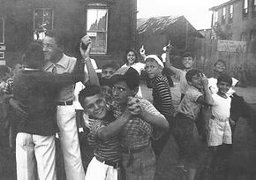 A group of children and a young woman outdoors in a neighborhood, some children smiling and playing, with houses in the background.