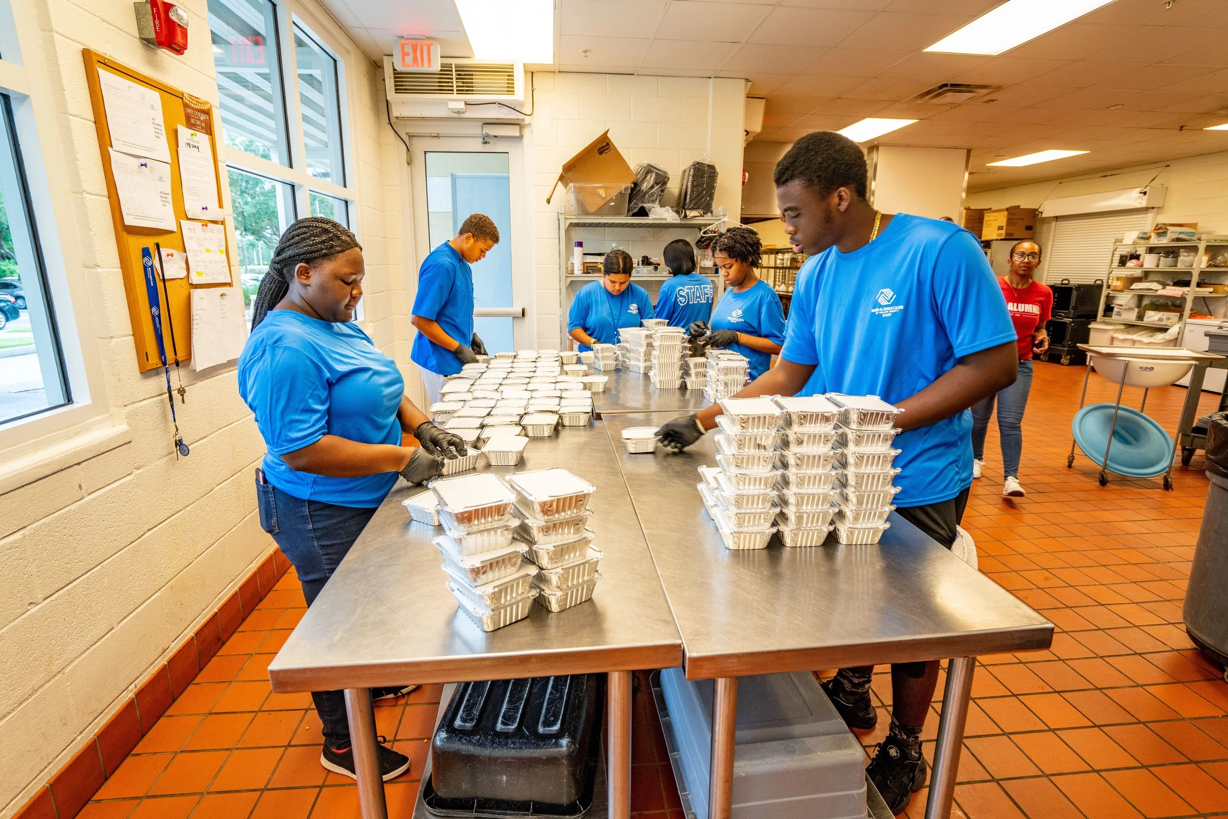Group of young volunteers in blue shirts packing food containers on a stainless steel table in a food distribution center.