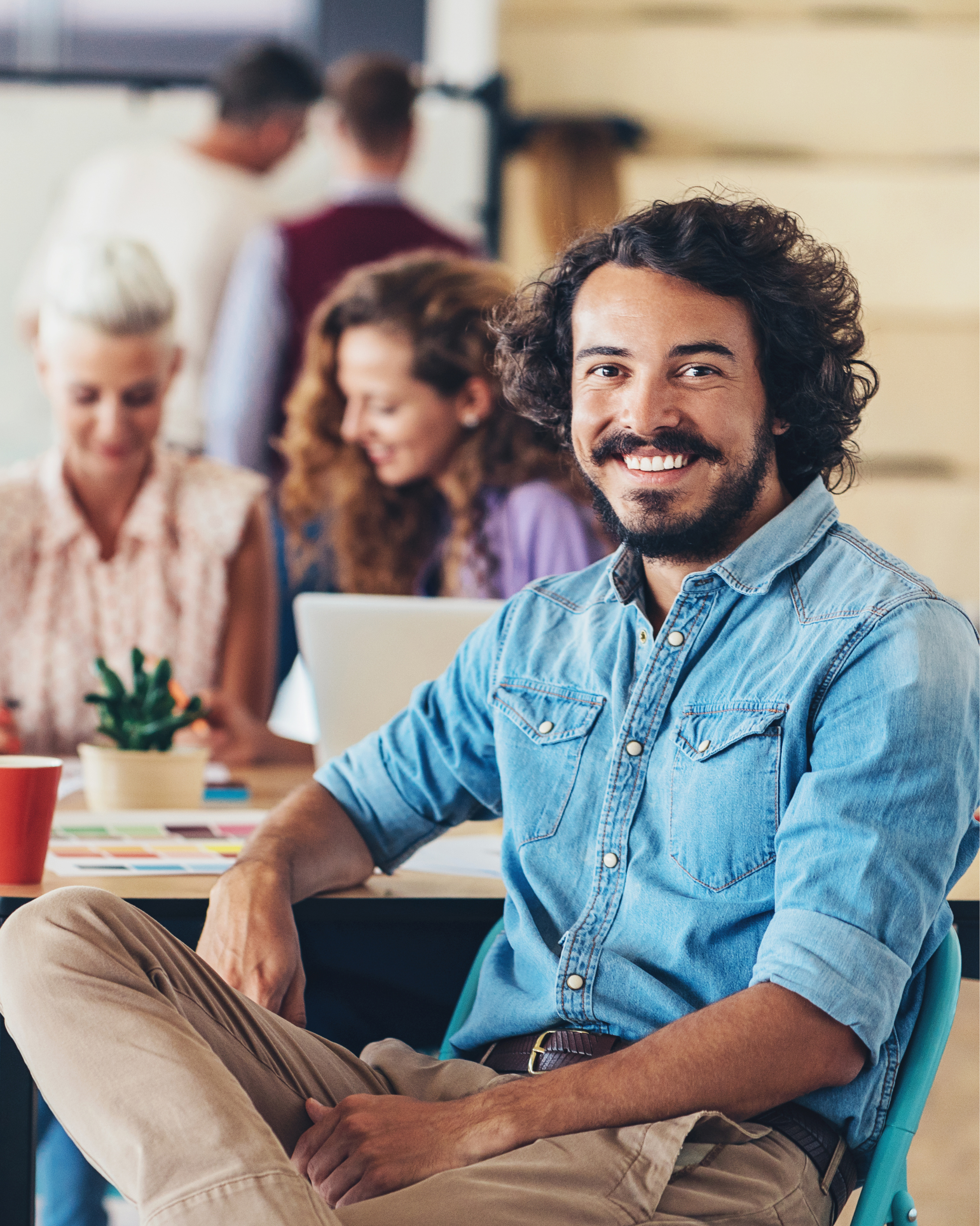 A smiling man with curly hair and a beard wearing a denim shirt, sitting in a chair in a bright office or classroom setting with a group of people working on laptops in the background.