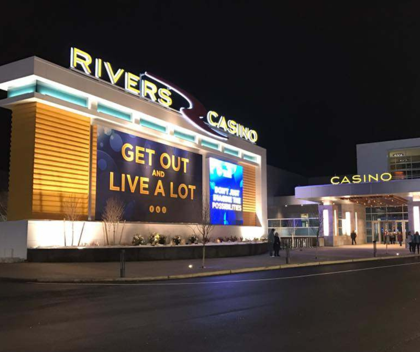 Night view of Rivers Casino, with bright signage and digital screens displaying messages, with a few people walking nearby.