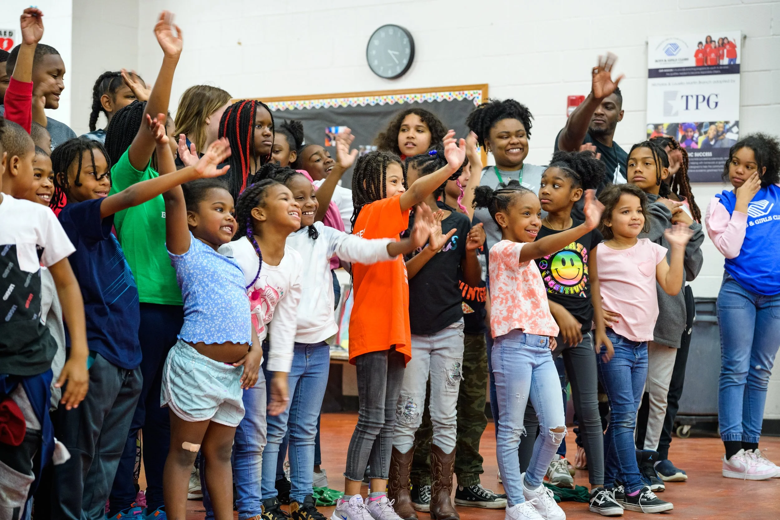 Group of children and adults smiling, cheering, and raising their hands in a school gymnasium or community center with a clock and a bulletin board in the background.