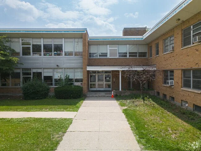 A two-story brick and glass school building with a concrete walkway leading to the entrance, small bushes, a leafless tree, and a traffic cone near the door.