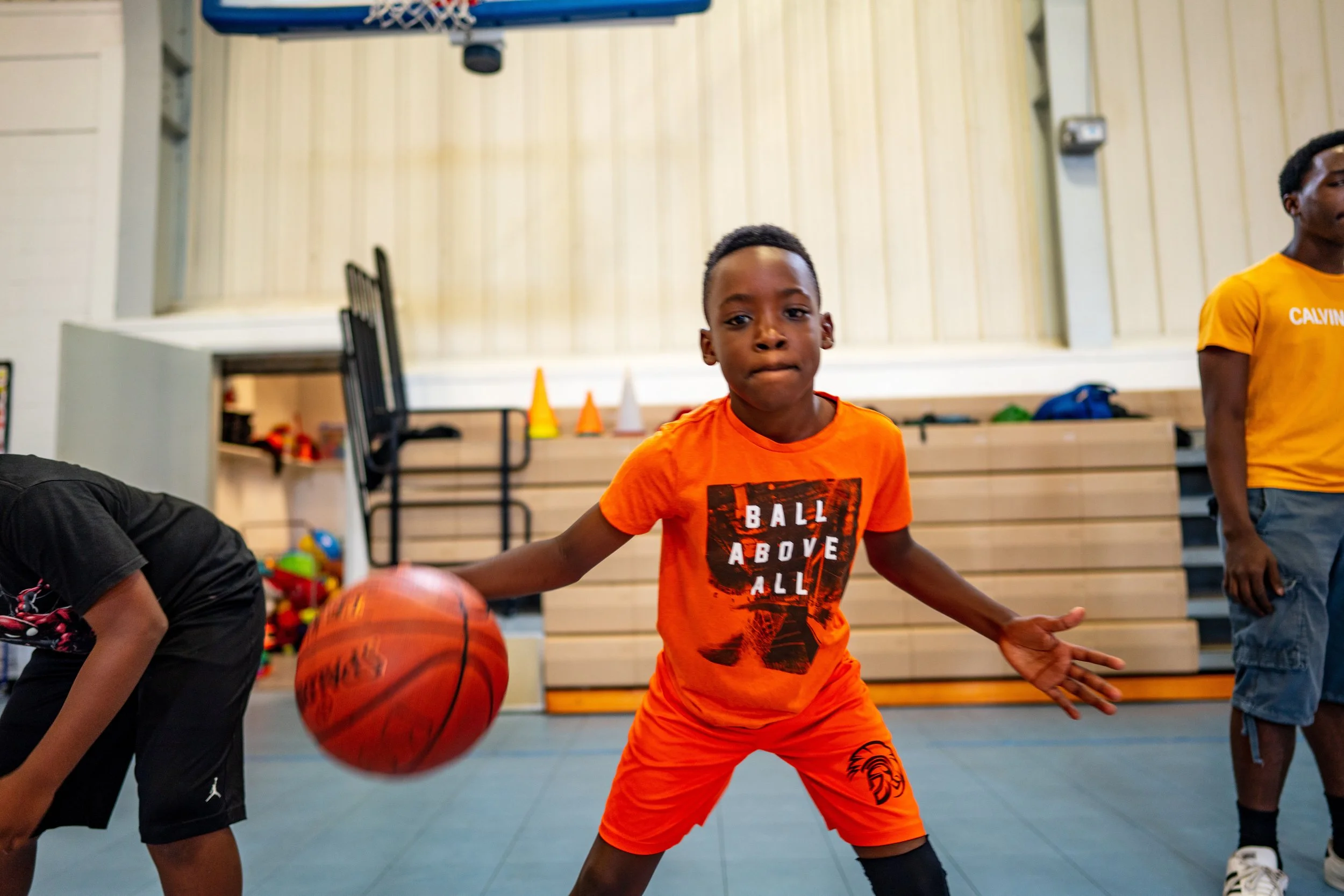 A young boy in an orange t-shirt and shorts is practicing basketball dribbling in a gymnasium, with other children nearby.