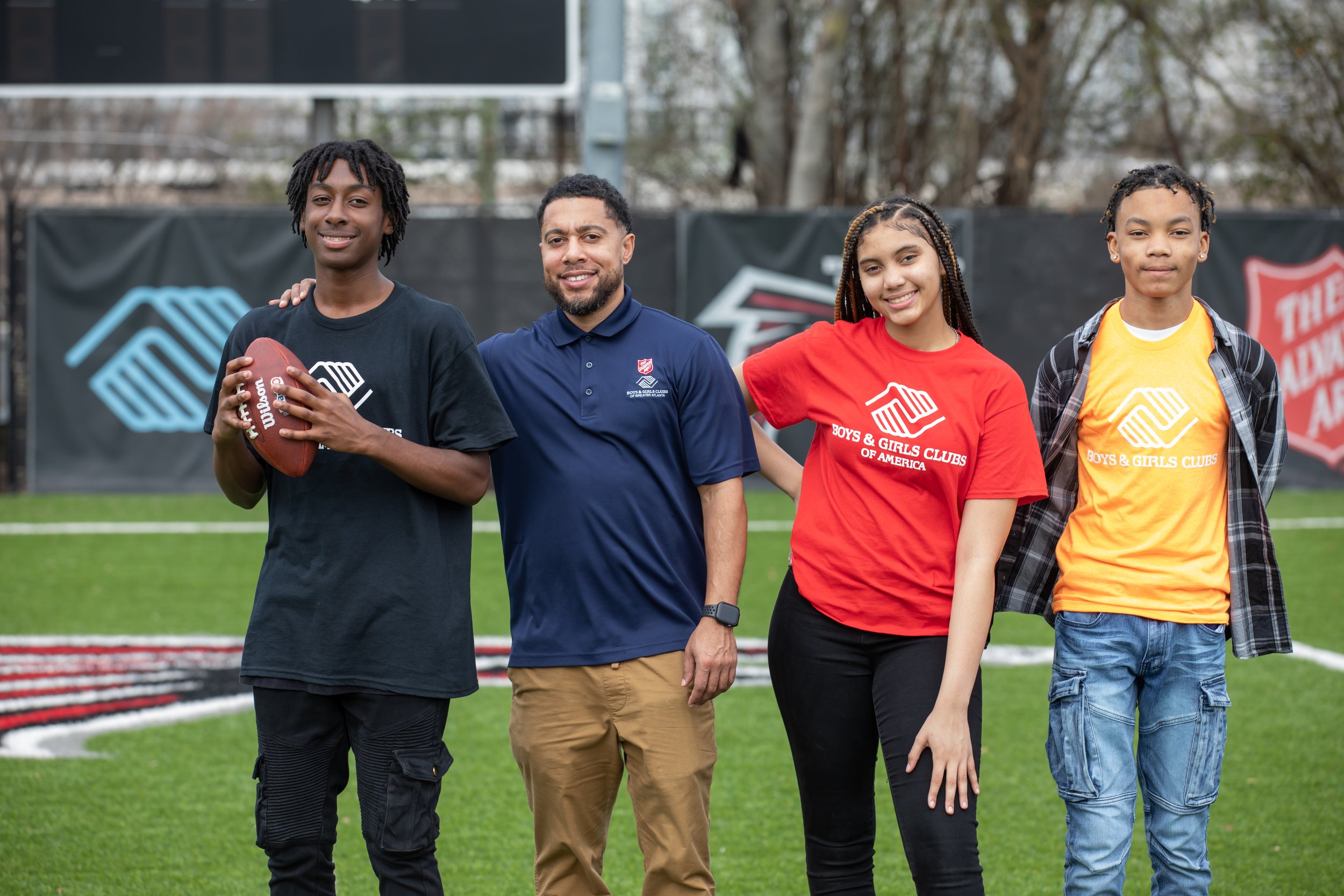 Four individuals, a young man holding a football and three others, standing on a football field with the Boys & Girls Clubs of America logos on their shirts, posing for a photo.