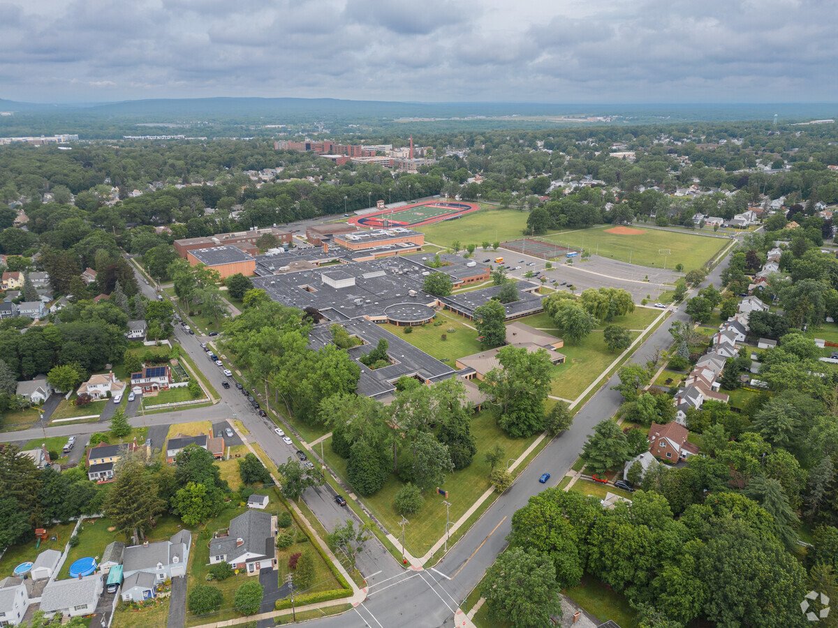 Aerial view of a school campus with buildings, parking lot, sports fields, and surrounding neighborhood with trees.