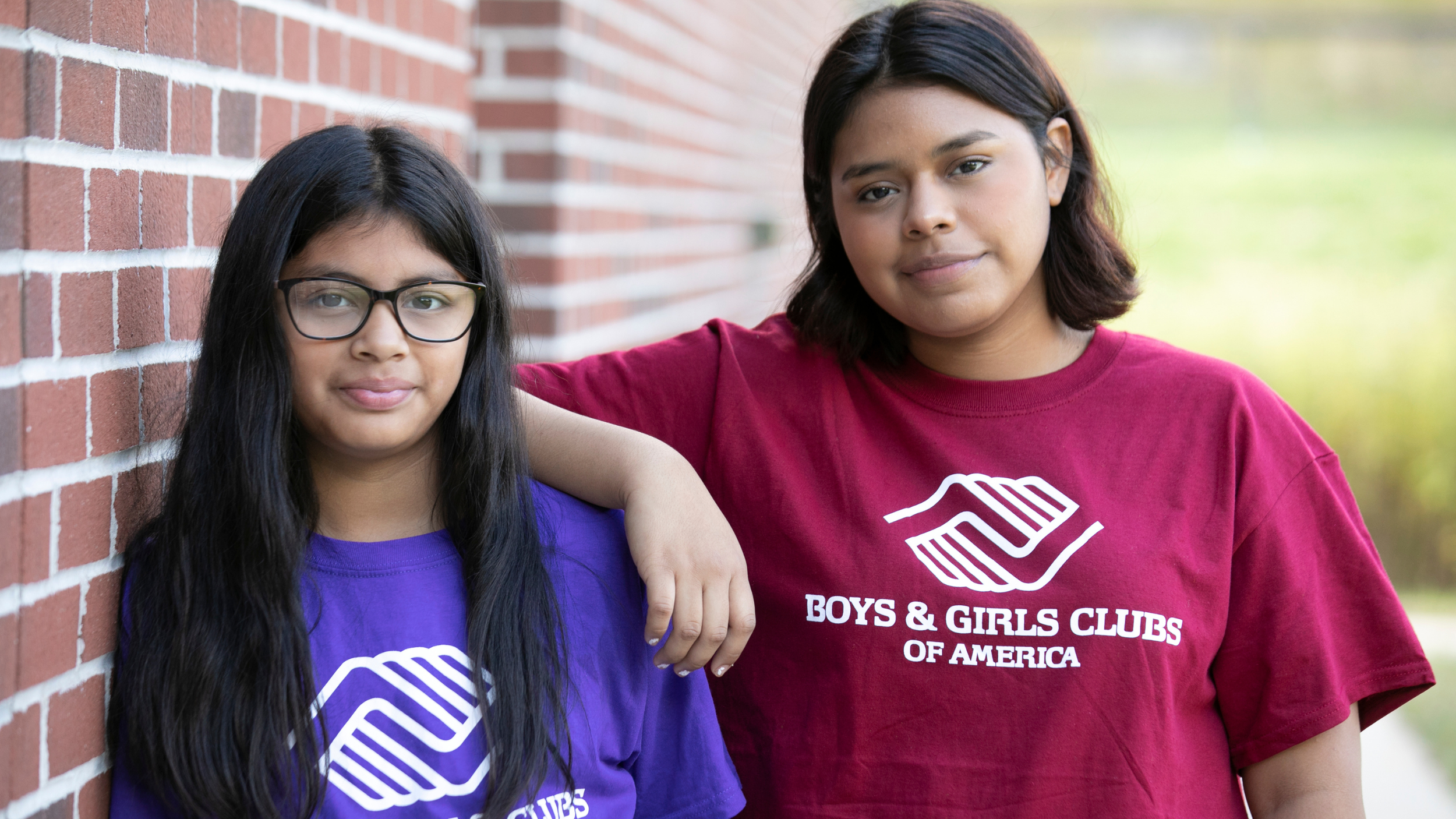 Two girls standing outside, one with glasses and long black hair, wearing a purple Boys & Girls Clubs of America t-shirt, and the other with short dark hair, wearing a red Boys & Girls Clubs of America t-shirt, leaning against a brick wall.