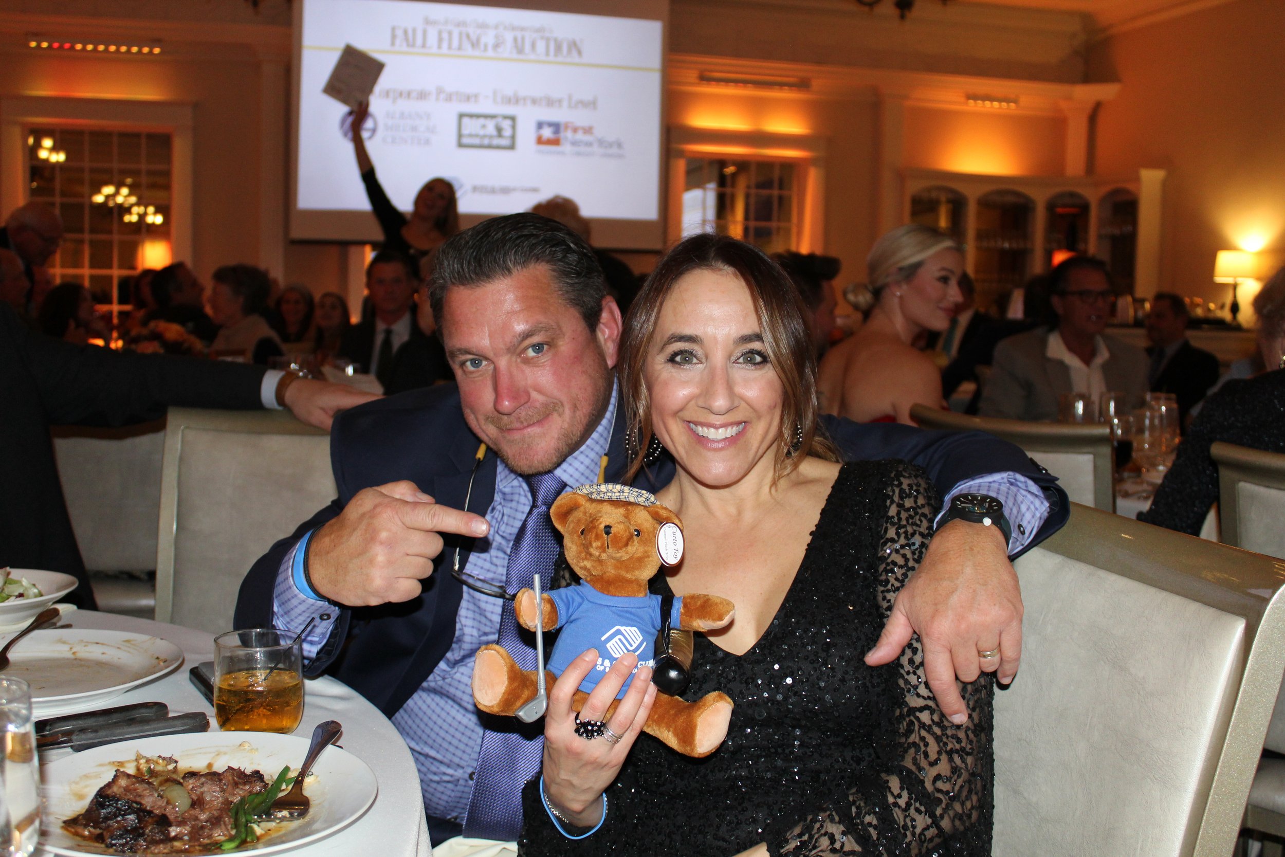 A man and a woman smiling and sitting together at a banquet or dinner event, holding a teddy bear dressed in a blue sports shirt, with people in the background and a large screen displaying a charity auction.