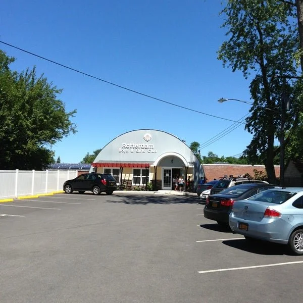 Parking lot in front of Rotocream Boy Scout clubhouse with several parked cars, white fence, and trees under a clear blue sky.