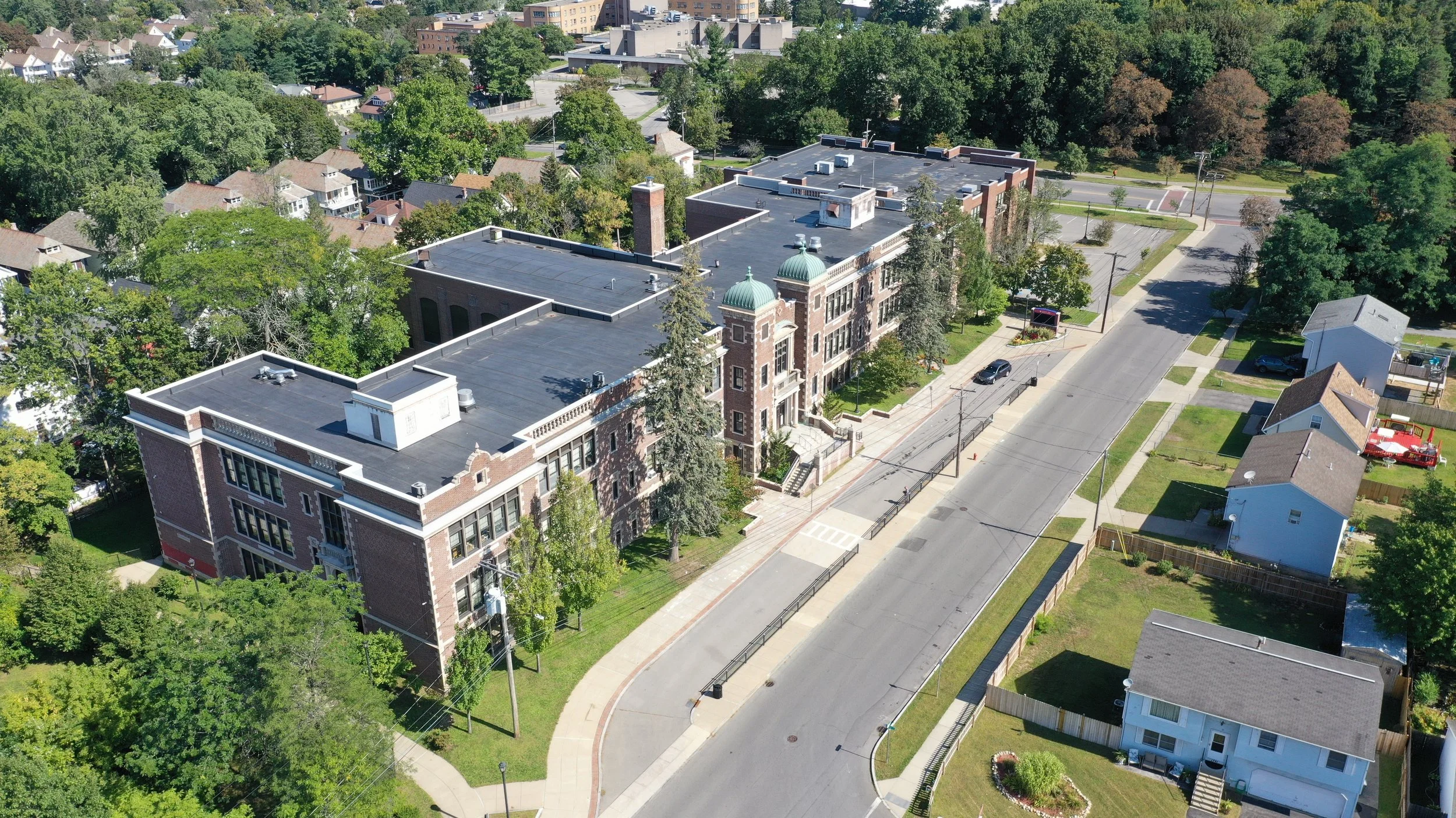 An aerial view of a historic brick building with green domed towers, surrounded by trees and residential houses on a sunny day.