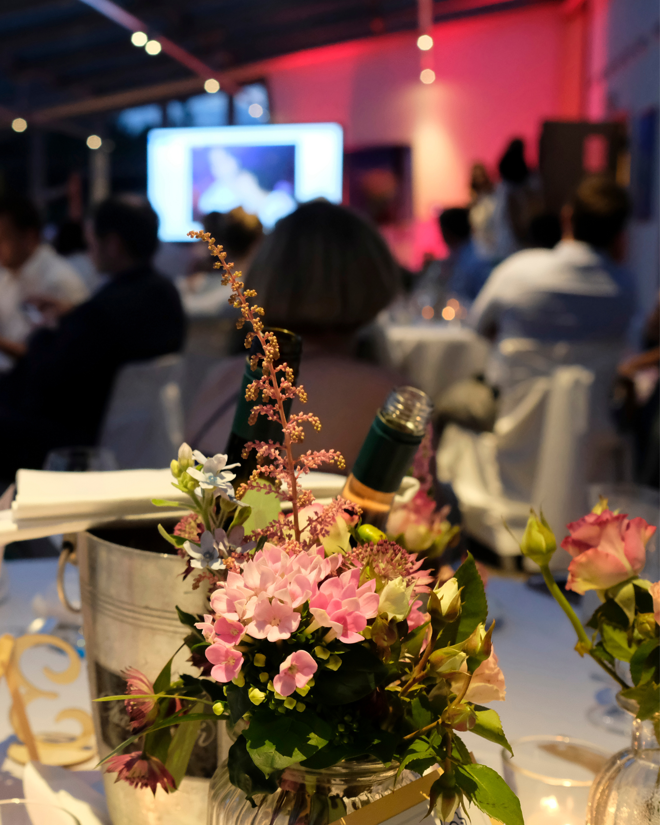 A table centerpiece with pink and white flowers, a wine bottle, and napkins at a formal event with people seated in the background, watching a presentation or performance on a screen.