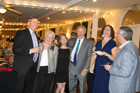 A group of six adults dressed in formal attire, smiling and enjoying a social event with string lights and decorated tables in the background.