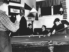 People playing pool in a recreational room, with one person taking a shot and others watching, in a black-and-white photo.