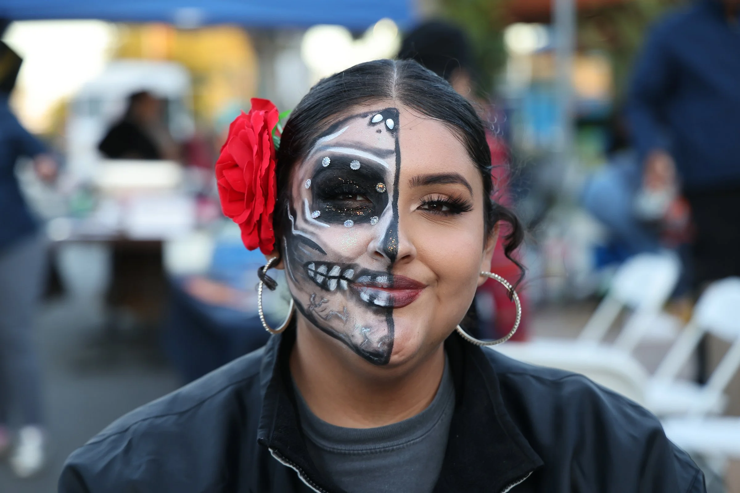 Young woman with half face painted as a skull, decorated with rhinestones, and a red flower in her hair, smiling at an outdoor event.