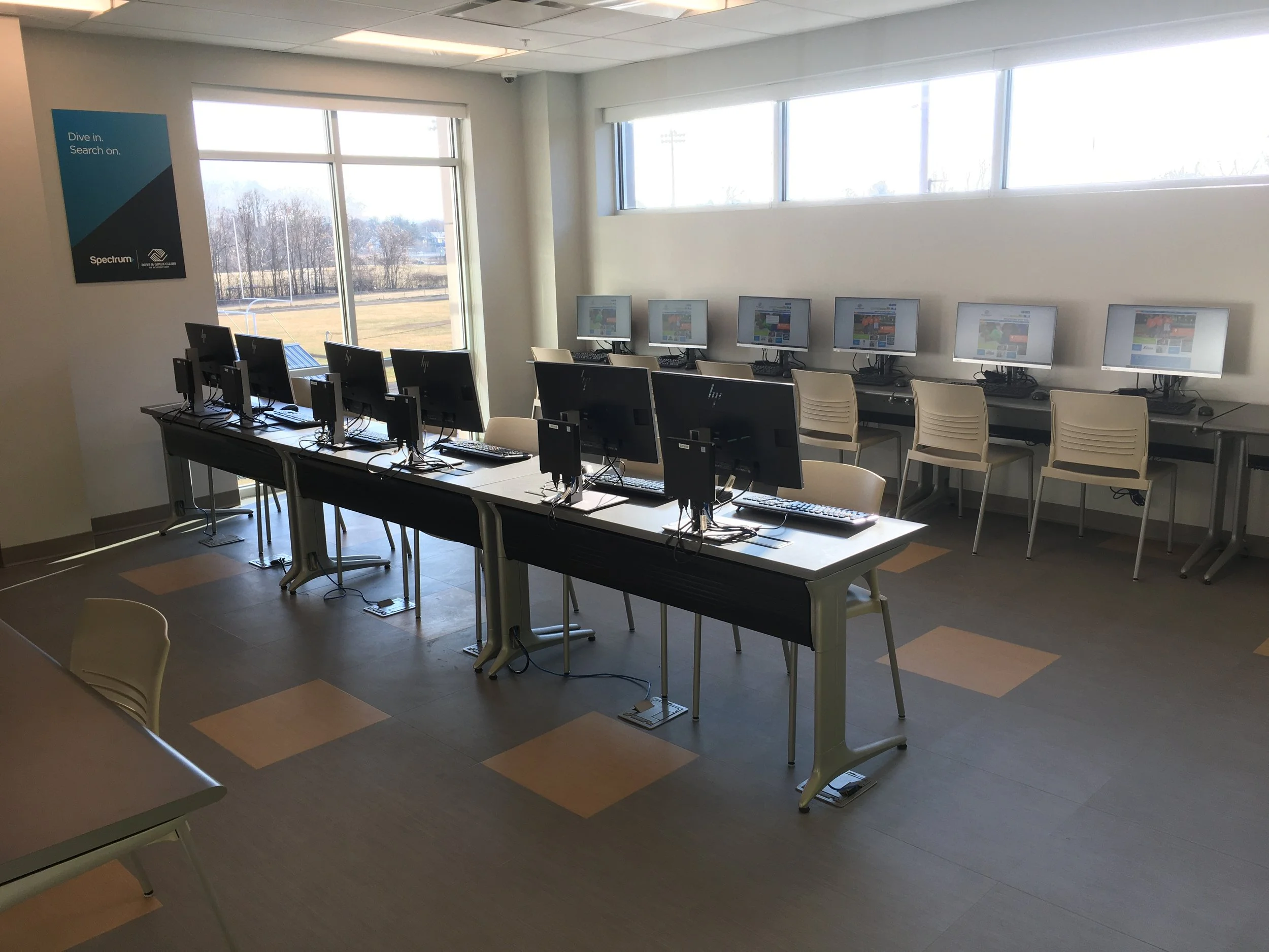 Computer lab with two rows of desktop computers, all turned on, in a room with large windows letting in natural light, and a poster on the wall.