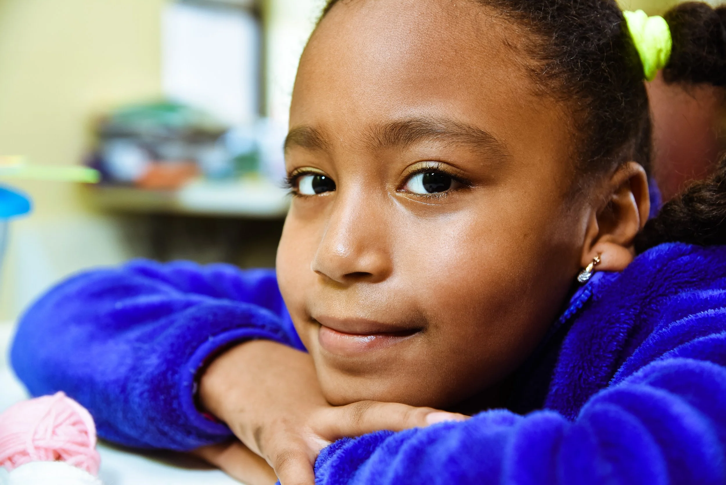 Close-up of a young girl with braided hair and earrings, leaning on her arms and looking at the camera with a gentle smile in a classroom setting.