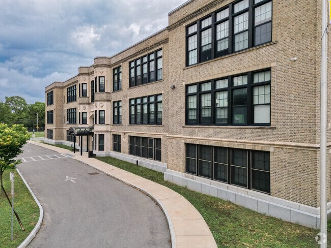 Exterior view of a multi-story brick building with large black-framed windows and a sidewalk leading to the entrance, with a curved road and some greenery in the surroundings.