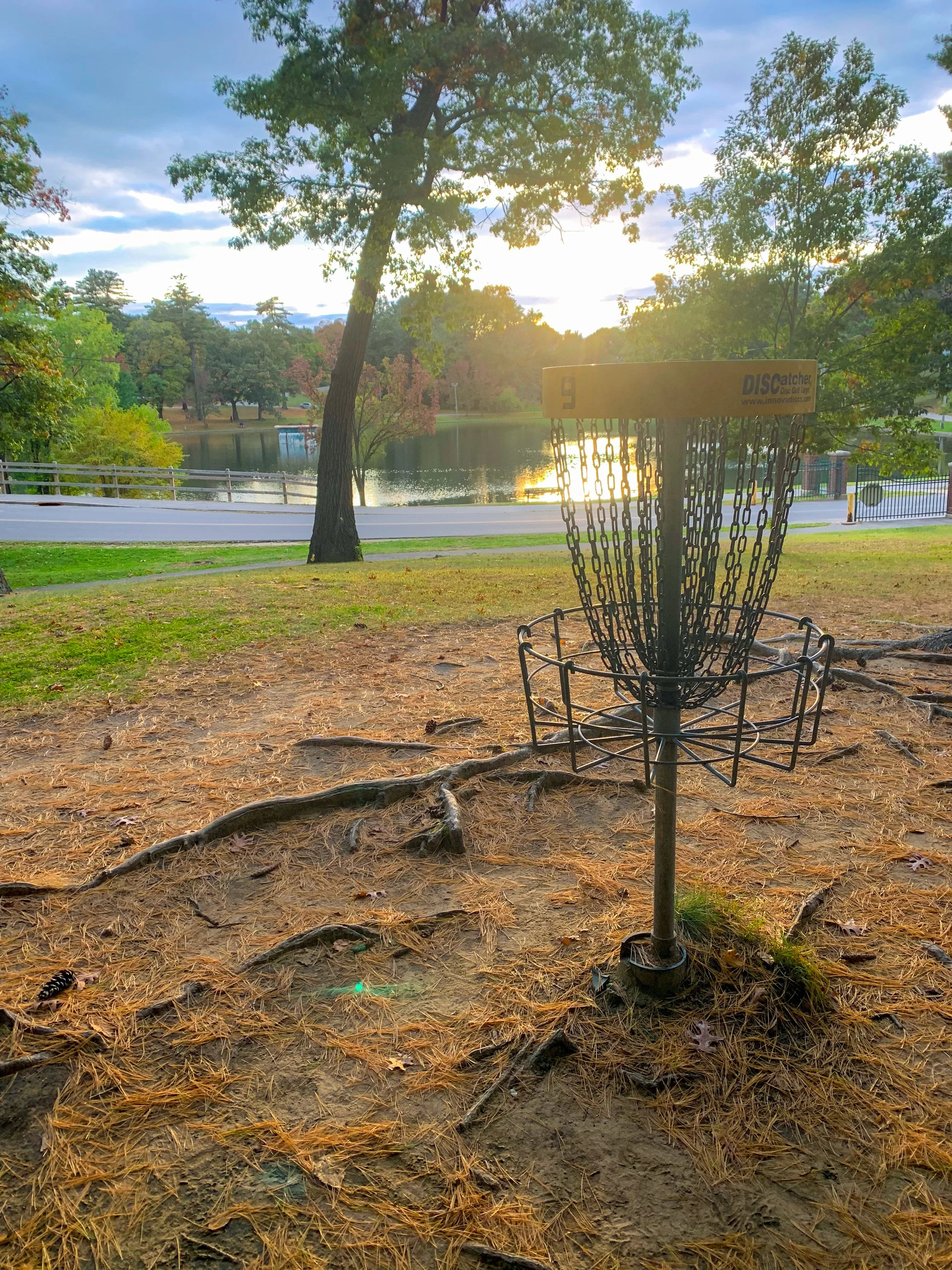 Disc golf basket near a lake with trees and a sunset in the background.