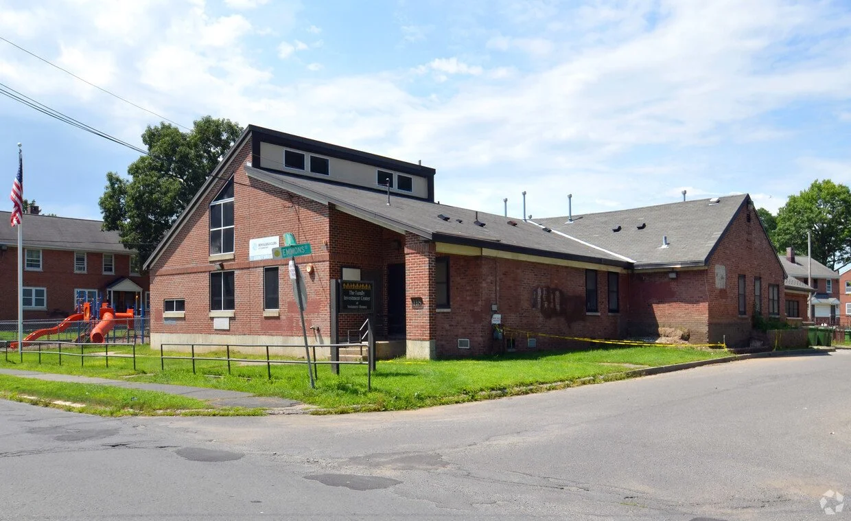 A red brick building on a street corner with a playground to the left and a flagpole with an American flag. The building has a sloped roof, several windows, and a small sign near the entrance. Yellow caution tape surrounds part of the building, suggesting ongoing construction or repairs.