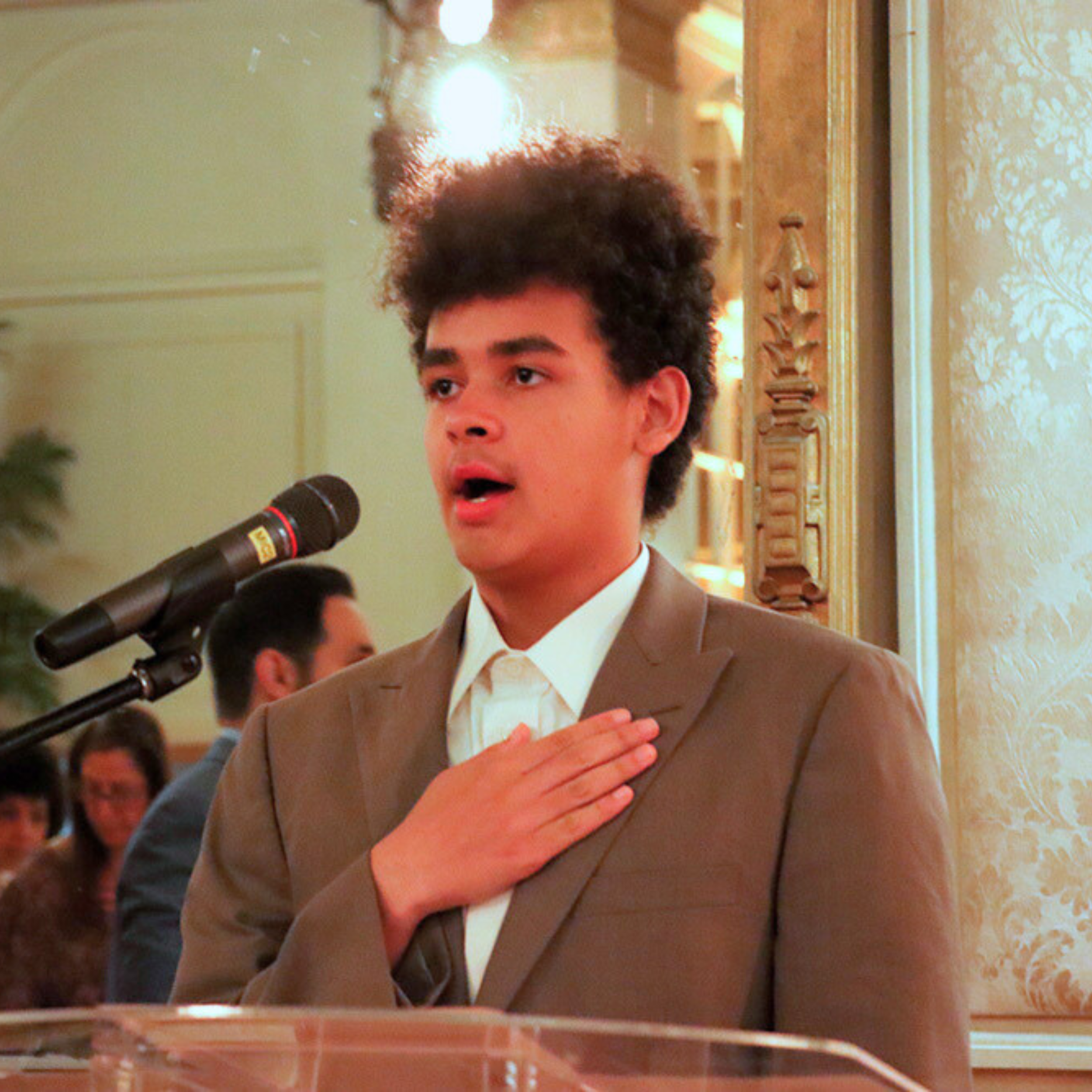 A young man with curly hair wearing a brown suit and white shirt is standing with his right hand on his chest, speaking into a microphone during a formal event.