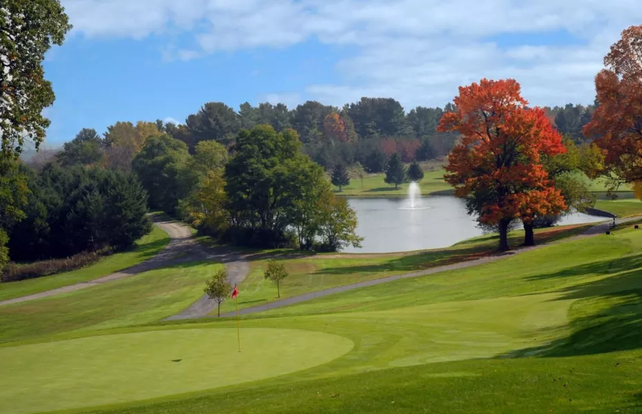A golf course with a green, fairway, a pond with a fountain, and trees with autumn foliage.