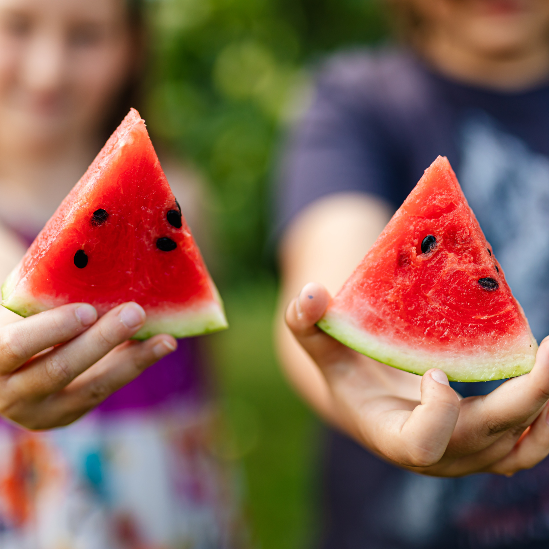 Two children holding slices of watermelon outdoors, blurred background with greenery.