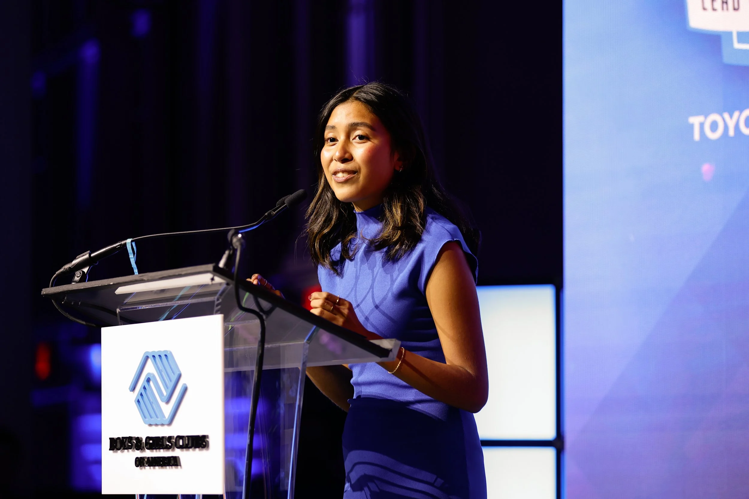 A woman in a blue dress speaking at a podium with a microphone during a conference.