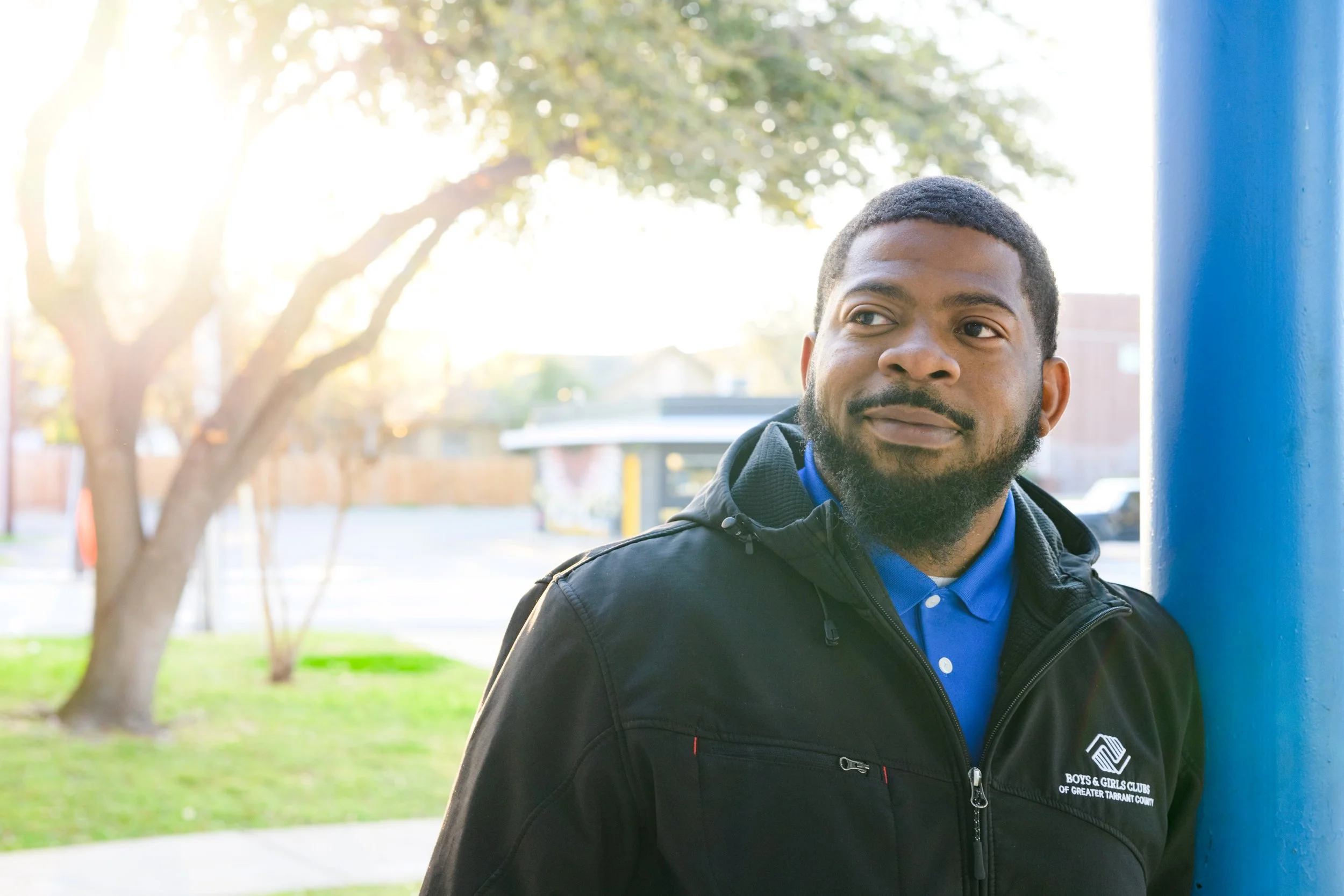 A man with a beard and mustache leaning against a blue pole outdoors on a sunny day, wearing a black jacket with a 'Boys & Girls Clubs' logo.