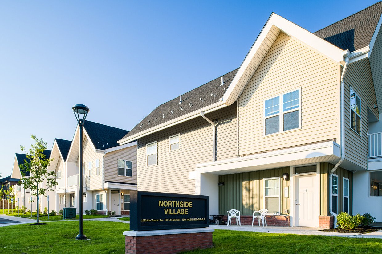 Residential apartment complex named Northside Village with multi-story houses, green lawn, trees, street lamp, and a sign showing the complex's name and address.