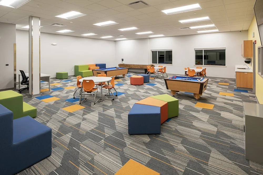 A spacious, modern recreational room with colorful furniture, including orange chairs, green and blue cushioned seats, and two pool tables. The room has a patterned carpet and large windows, with a wall-mounted TV and a small kitchenette in the corne