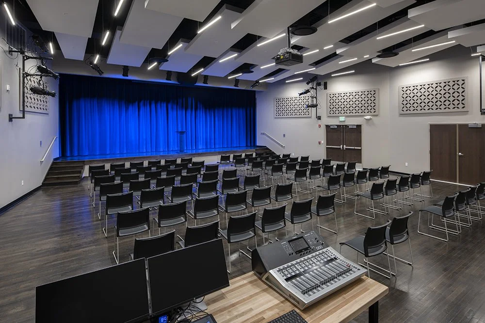 Empty auditorium with a stage, blue curtains, rows of black chairs, audio and lighting equipment, and a sound console.