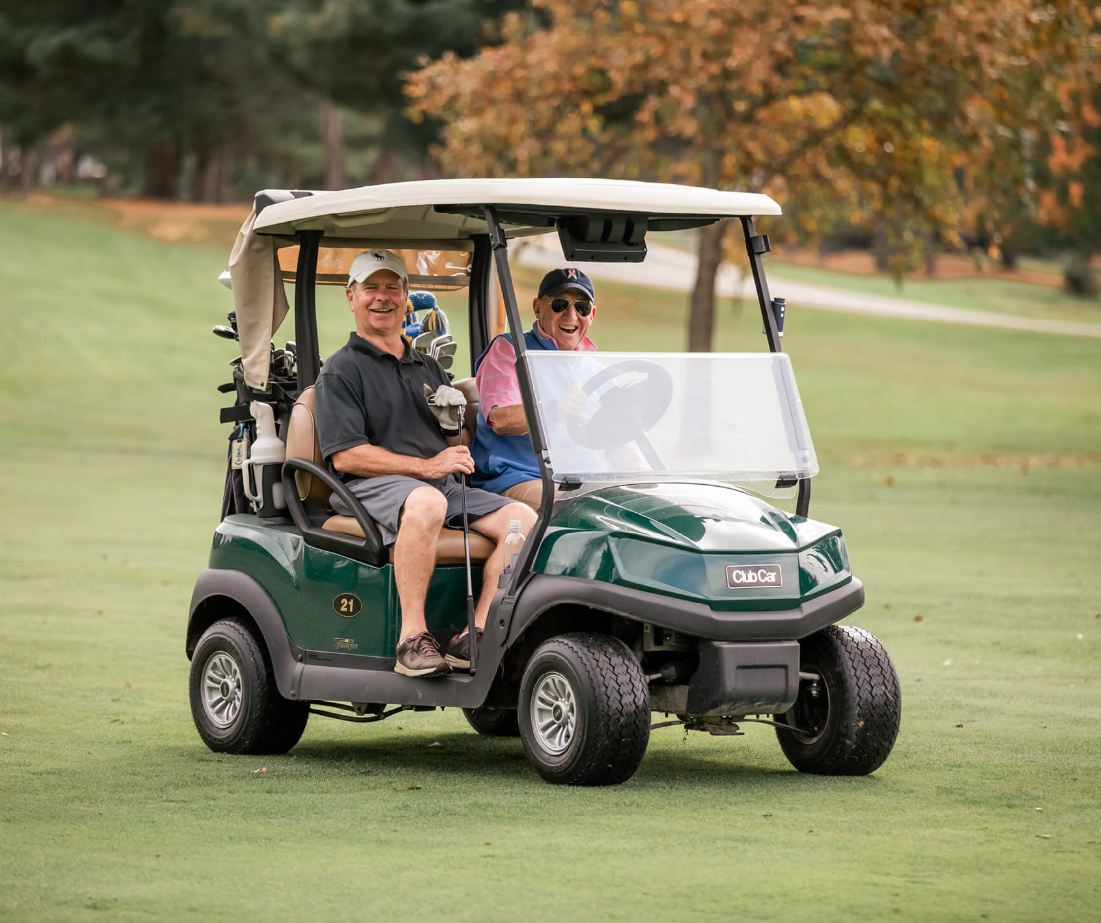 Two men sitting in a golf cart on a golf course, smiling and looking cheerful. One is in the driver's seat holding the steering wheel, wearing sunglasses and a navy cap. The other man sits beside him, holding a golf club and wearing a light-colored cap. The golf cart is on the green, with trees in the background showing fall foliage.