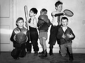 Five children holding sports equipment, including basketballs, a boxing glove, and a tennis racket, in a gym setting.
