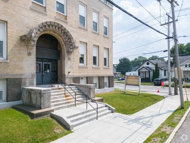 A beige brick building with an arched entrance and steps, situated on a sidewalk with grass and a street with utility poles and houses in the background.