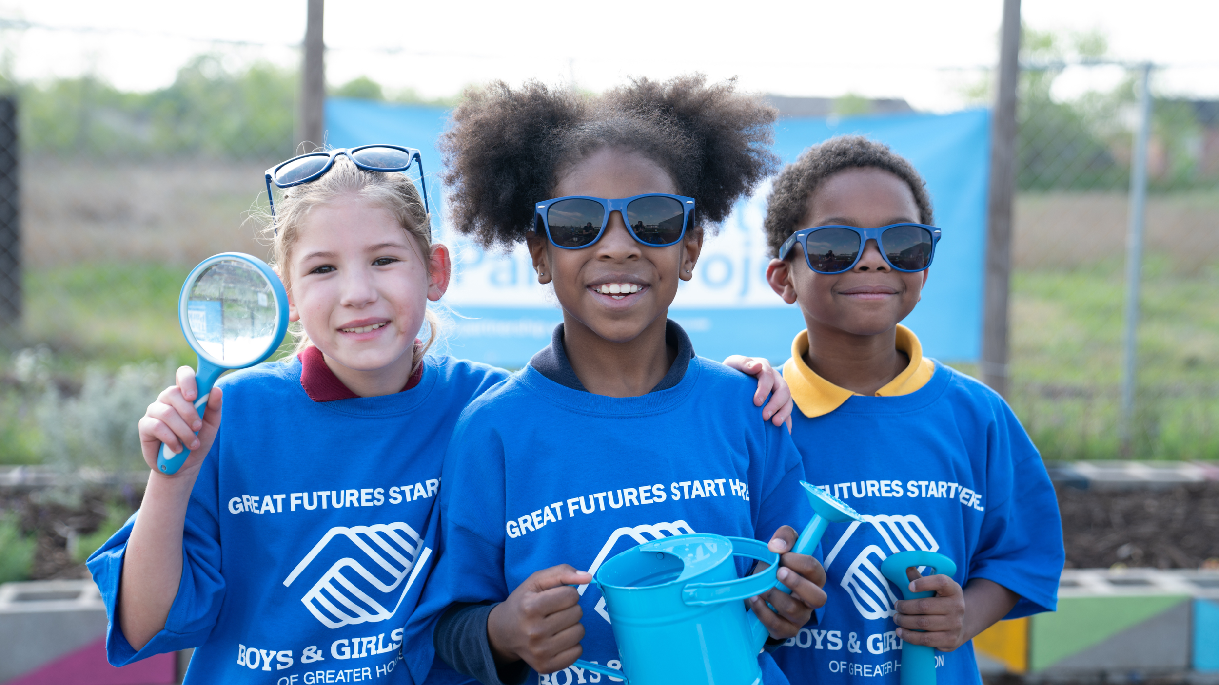 Three children wearing matching blue shirts with the words "Great Futures Start Here Boys & Girls" posing outdoors together, smiling, with one holding a magnifying glass, another holding a blue watering can, and the third with a small gardening tool,
