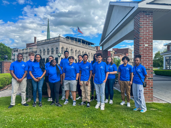 Group of eleven young people standing outdoors on grass in front of a building and pavilion, all wearing blue shirts. Buildings and a flag are visible in the background.