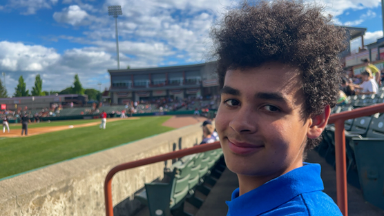 A young man with curly hair smiling at a baseball game at a stadium with blue skies and some clouds.
