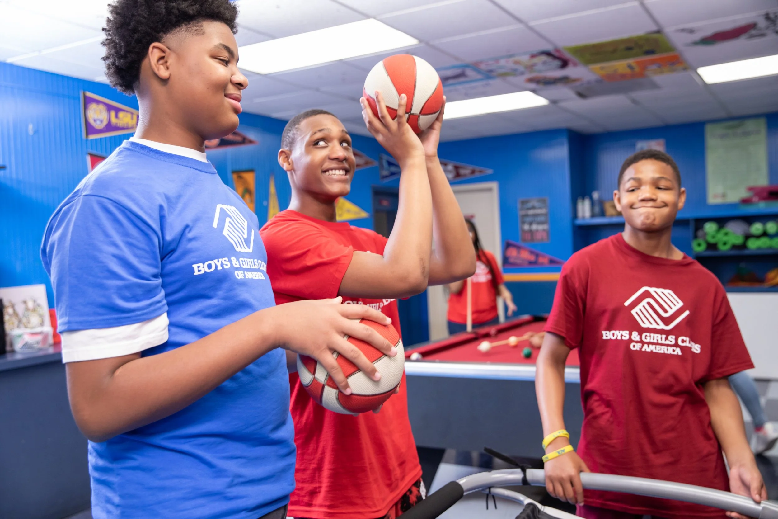 Three boys at an arcade or recreational center, one holding a basketball, another smiling with a pool cue, and the third standing with a smile, all wearing Boys & Girls Clubs of America shirts.