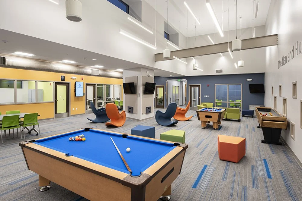 Modern indoor recreation room with pool table, foosball table, lounge chairs, and a small dining area, decorated with colorful furniture and large windows.
