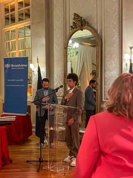 A woman in a brown suit standing at a microphone in a decorated room with a large mirror and ornate wall decorations, while a man assists her. Another woman in a pink outfit is partially visible in the foreground. A blue banner with the text 'Broadvi