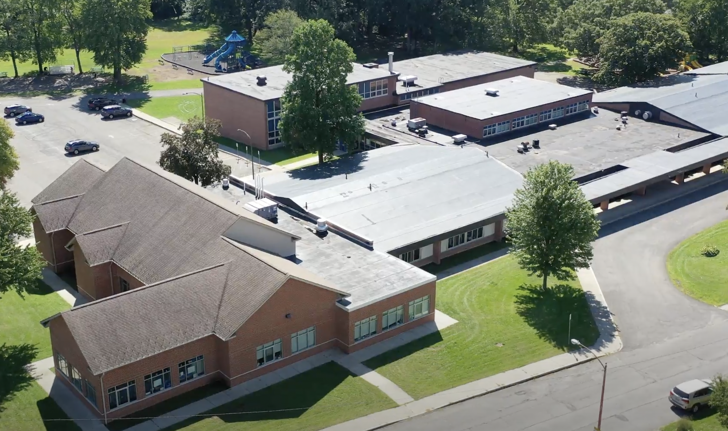 An aerial view of a school complex with multiple single and multi-story brick buildings, a parking lot with several cars, a playground with a slide, and surrounding green trees and grass.