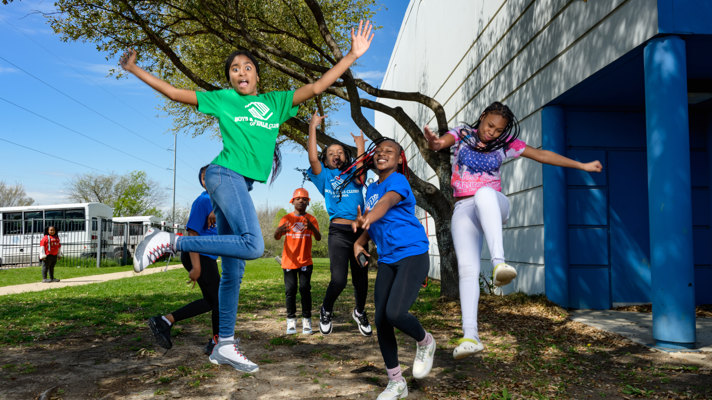 Group of children playing and jumping outdoors under a tree, some wearing Boys & Girls Clubs T-shirts.