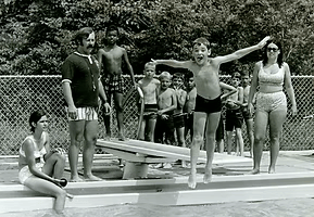 A group of children and teenagers at a swimming pool. One boy is jumping into the water with arms outstretched, while others watch nearby.