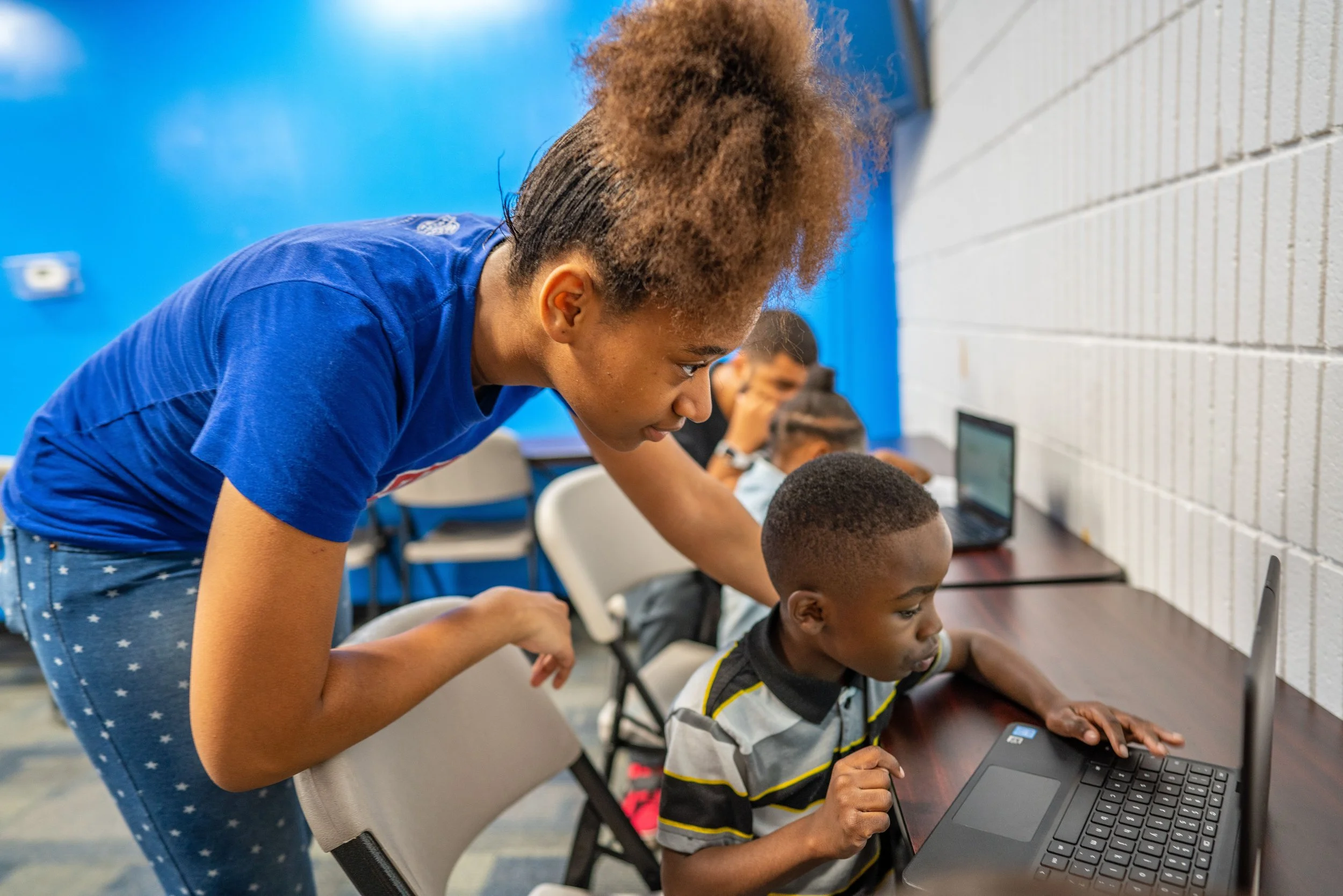 A young woman leaning over a boy working on a laptop in a classroom setting with other children working on computers in the background.