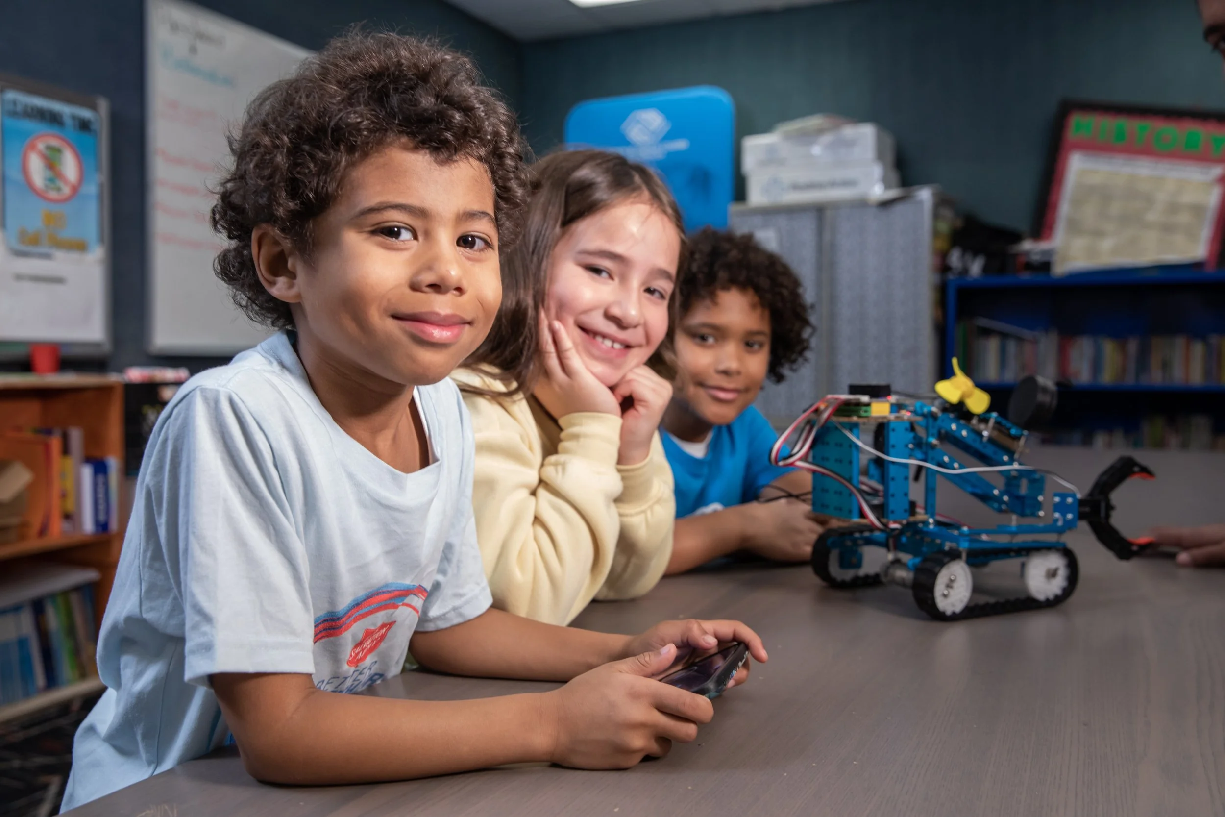 Three children sitting at a table with a robotic vehicle, smiling at the camera, in a classroom or library setting.